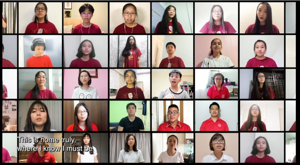 A virtual choir of students in red shirts singing, with subtitle text reading "This is home truly, where I know I must be."